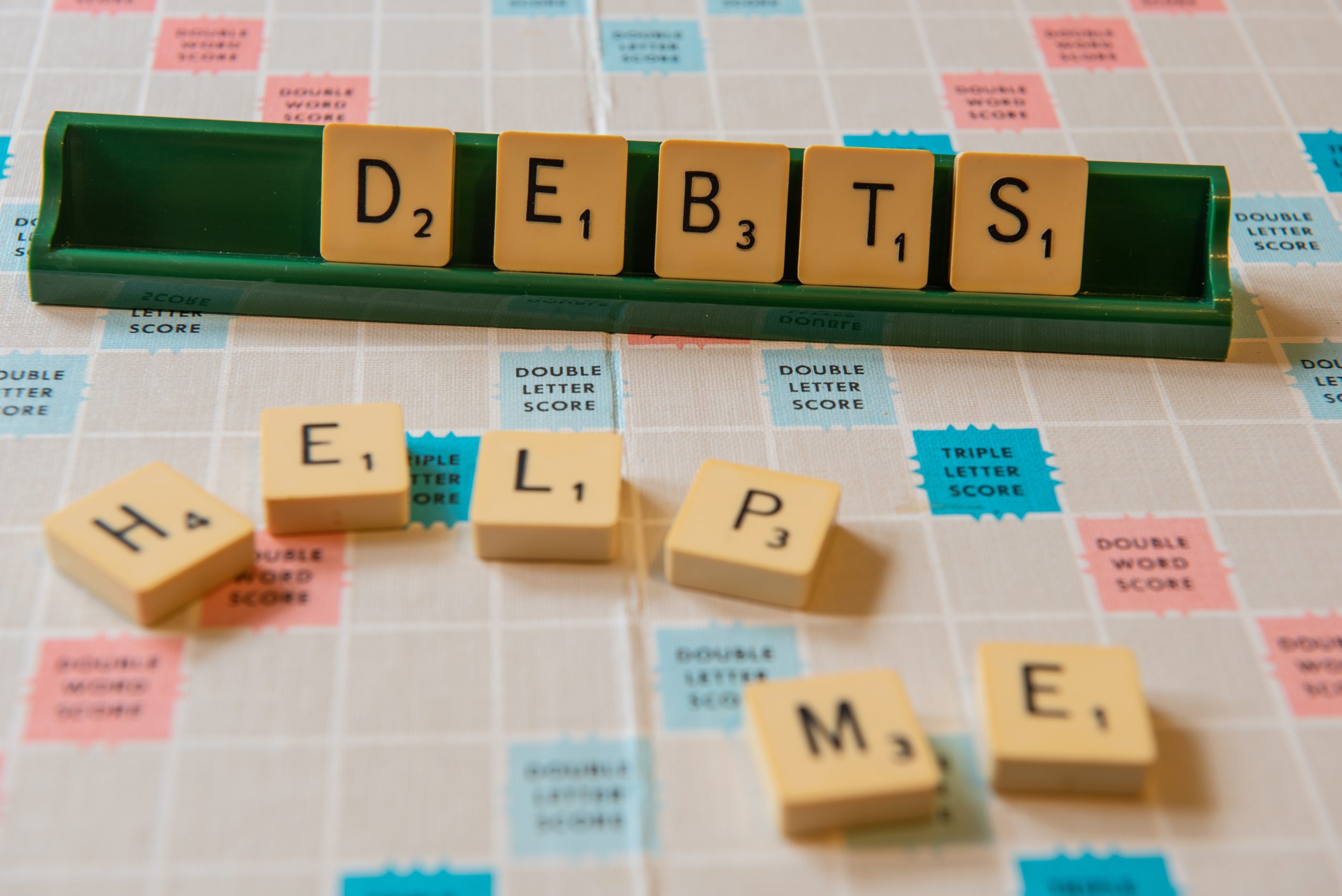 Close-up view of the words "DEBTS HELP ME" arranged on a scramble board with glowing lights in the background, symbolizing the urgent need for a debt consolidation plan in Singapore.