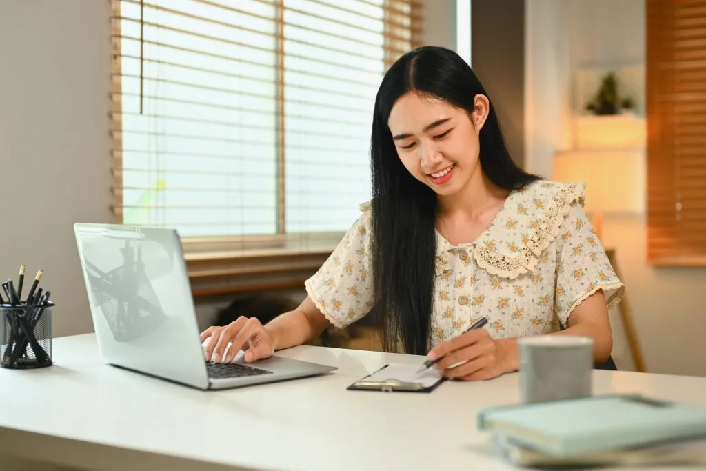 Pretty young Asian woman using laptop and making notes browsing